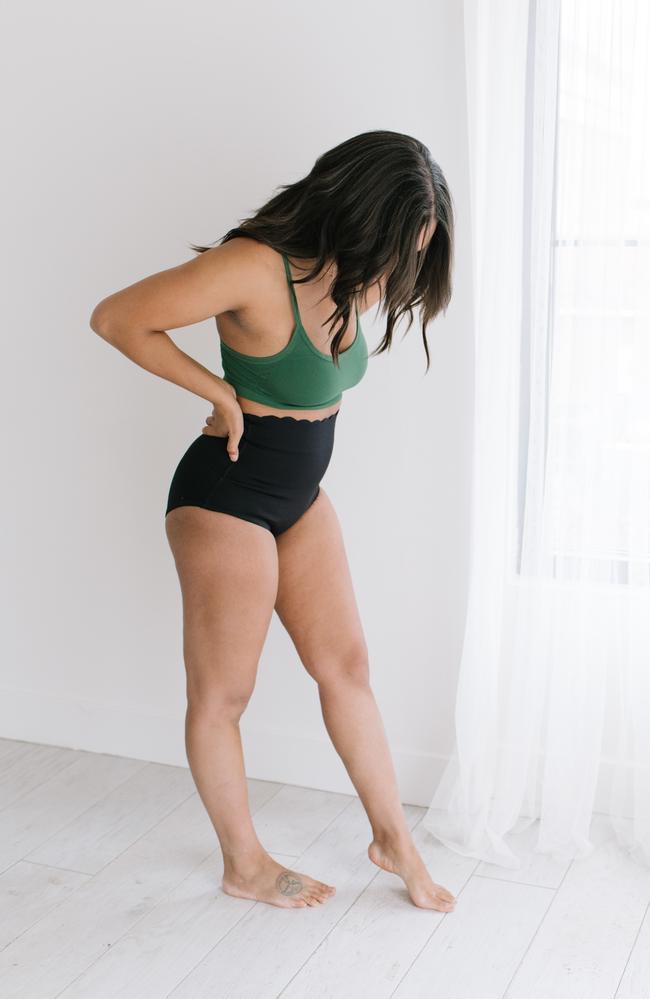 Woman standing in front of window wearing high waisted black underwear and green bralette